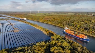 Container ship and car ferry navigate the Kiel Canal alongside vast solar panel fields and wind turbines, set against a backdrop of forest.
