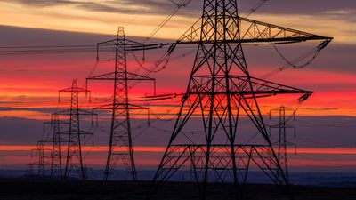 A breathtaking sunset paints the sky with vivid hues of orange, purple, and red, forming a dramatic backdrop for the imposing silhouettes of electricity pylons. These towering structures, carrying power lines, stand as symbols of modern energy infrastructure and the industrial landscape, captured in a moment of natural beauty and technological might.