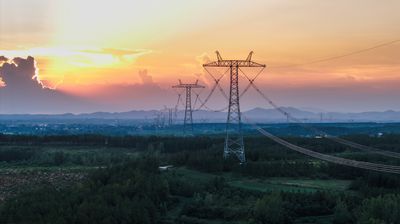 A beautiful sunset scene featuring high-voltage transmission towers standing in a vast field with greenery and distant mountains.