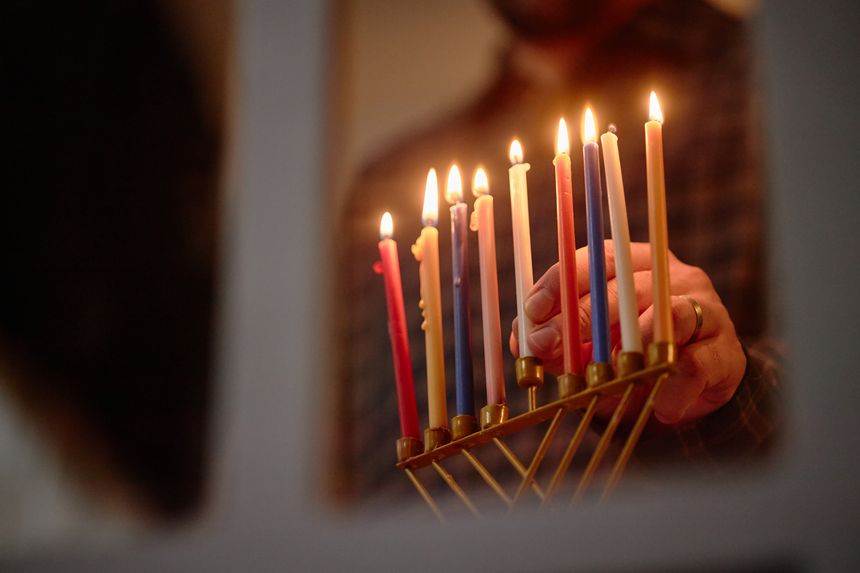 Man lighting candles on menorah during Hanukkah celebration, hand holding match while menorah with colorful candles standing on table, focus on ritual action