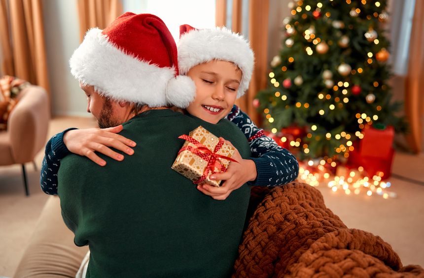 A happy boy hugs and holds a gift from his dad while wearing a Santa hat on Christmas. Its an honest and joyful family moment, showcasing the love of children for their parents.