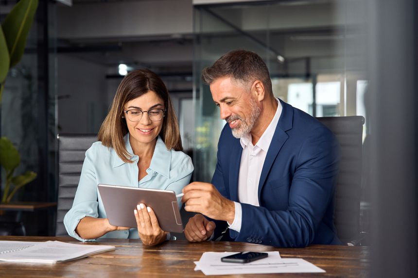 Team of diverse partners mature Latin business man and European business woman discussing project on tablet sitting at table in office. Two colleagues of professional business people working together.