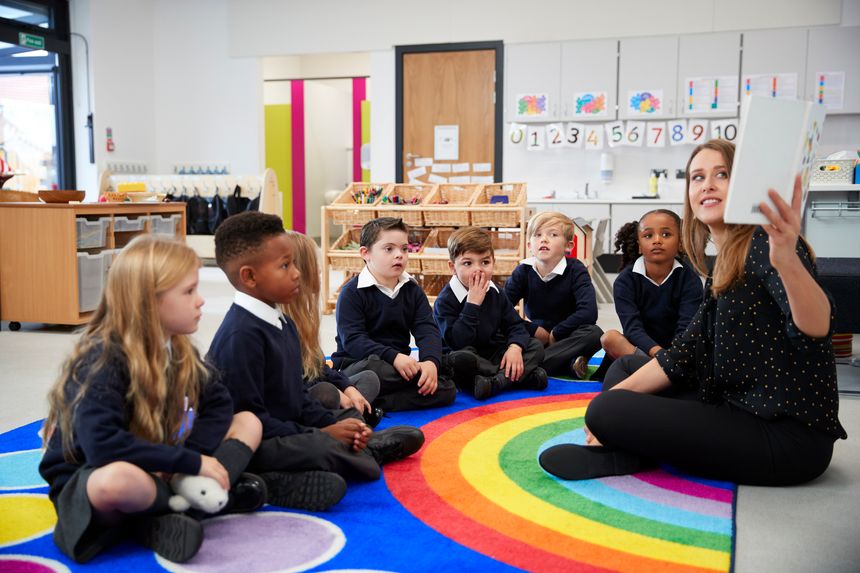 Female teacher holding up a book in front of her class of elementary school kids sitting on the floor in a classroom, side view
