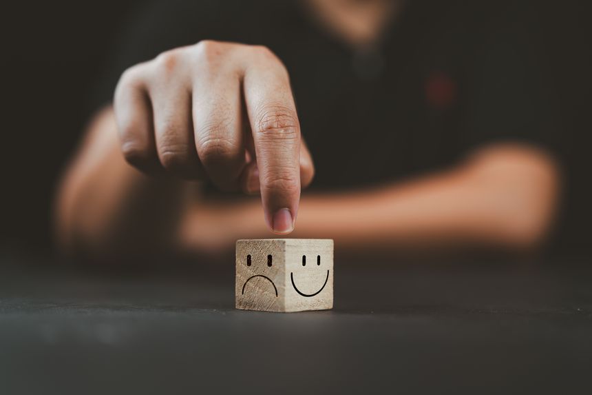 Man hand points on a wooden cube with happy smile face on bright side and unhappy face on dark side of wooden block cube for a positive mindset selection. Emotional state and mental health concept.