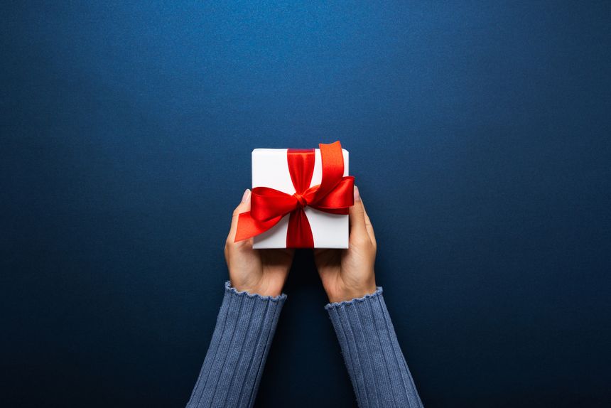 Flat lay showing the gesture of giving. A pair of hands, sleeves visible, carefully holding a single, neatly wrapped white gift box with a bright red bow, isolated against a minimalist dark blue backdrop. This image captures the essence of exchange, gratitude, and holiday giving. Perfect for illustrating concepts related to birthdays, Christmas, customer loyalty, or delivery services and special occasions.
