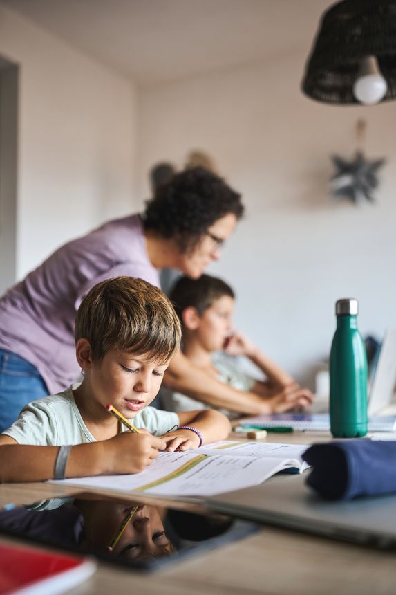 Homeschooling at home with children studying, boy writing in workbook while sibling uses laptop and mother supports learning indoors