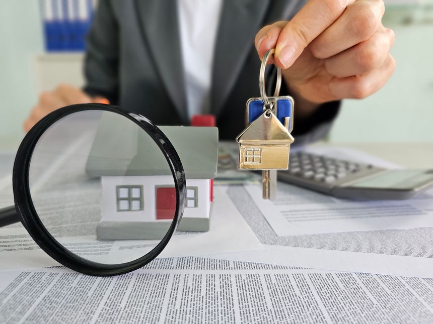 A person holds a set of house keys above a miniature house model while paperwork, a magnifying glass