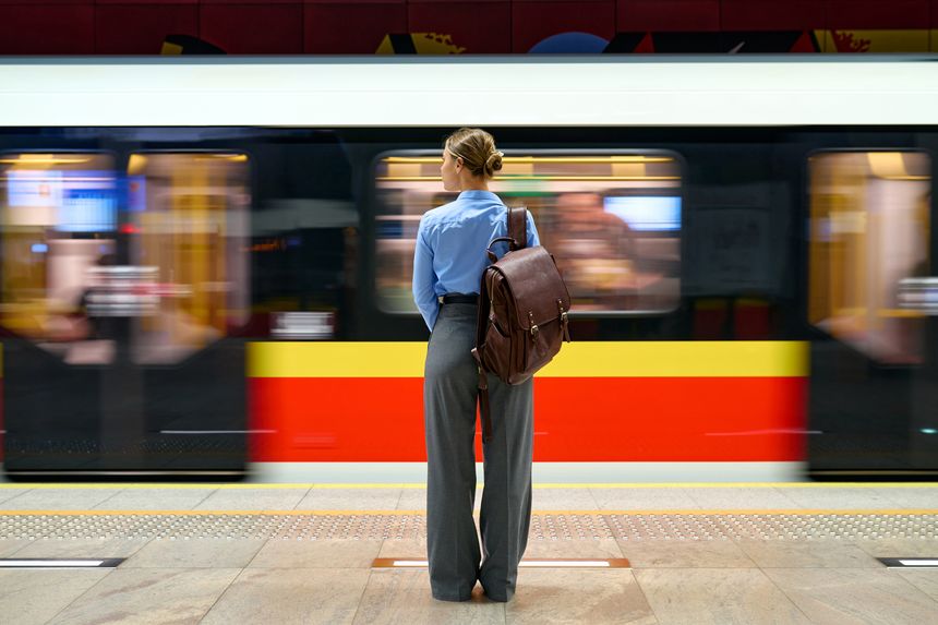Back view of young woman with brown leather backpack standing at subway platform, watching a fast-moving metro train pass by. Urban commuting, city life, and public transport theme.