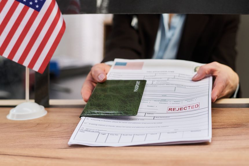 Caucasian middle aged woman handing over passport and visa application form with rejected stamp at visa center counter, American flag visible on desk, close up of hands and documents