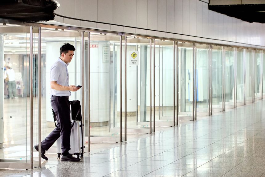 A Japanese businessman in his 30s walking through an underground walkway while holding a suitcase and using his smartphone. He is dressed in a suit and traveling for a business trip.