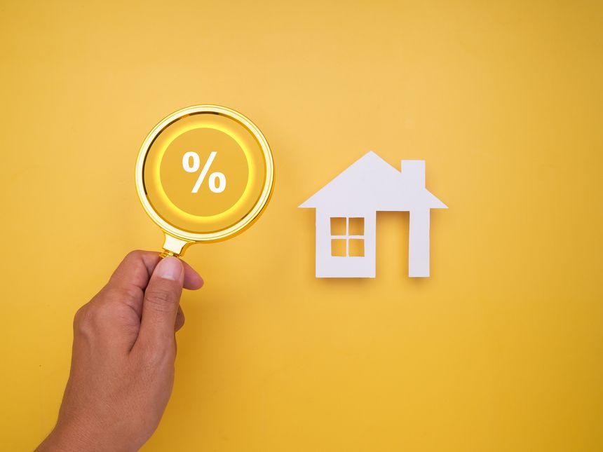 Close-up on home buying and financial analysis theme showing percentage symbol magnified near house shape, illustrating real estate costs and loan comparisons.