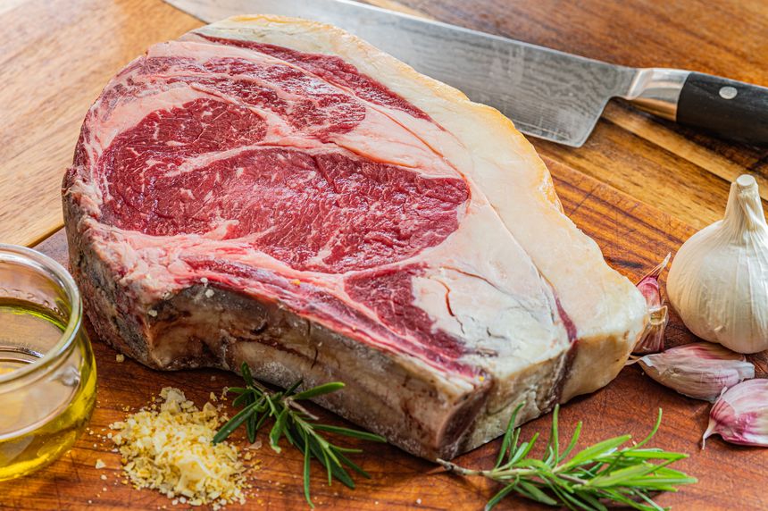 Aged vaca rubia gallega steak, ready to be grilled with garlic, rosemary, salt flakes and extra virgin olive oil and kitchen knife on wooden table