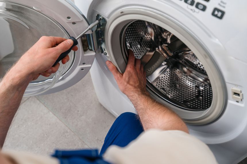 Cropped shot of a technician holding screwdriver tool, fixing door and working on repairing a broken washing machine in a modern laundry room. Residential apartment, maintenance service