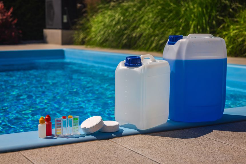 Set of pool maintenance supplies including liquid chemical canisters, chlorine tablets, and a test kit with dropper bottles placed by the pool on a sunny day. High quality photo