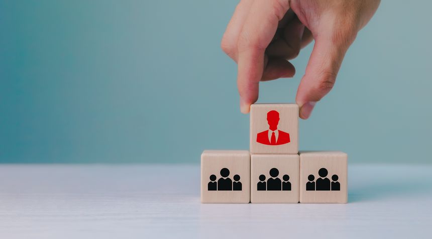 A hand places a wooden block with a red leader icon. Figure above three blocks showing groups of people. Organization and team structure symbolized with cubes, leadership organization hierarchy.