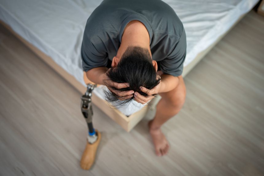Asian depressed young man with prosthetic leg sitting on bed in bedroom. Attractive male amputee wearing a leg brace feeling stress and upset with emotional challenges during recovery in cozy house.