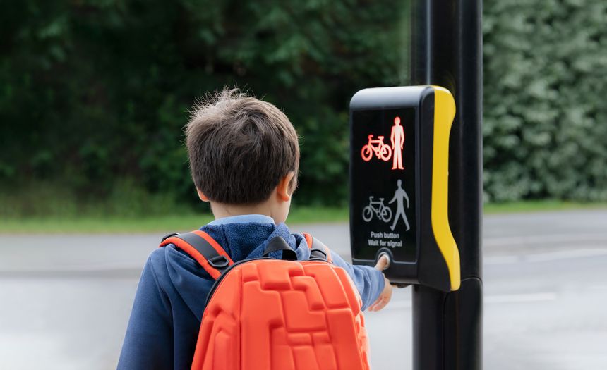 Rear view portrait School kid pressing a button at traffic lights on pedestrian crossing on way to school. Child boy with backpack using traffic signal controlled pedestrian facilities for crossing road.