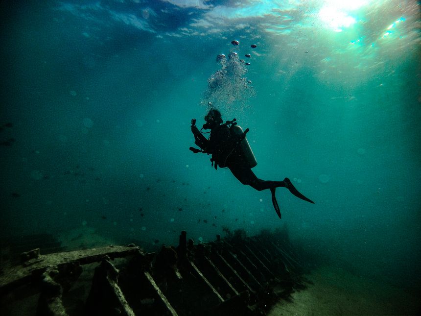 A lone scuba diver hovers above the skeletal remains of a sunken shipwreck, silhouetted against the deep blue ocean. Bubbles rise to the surface as sunlight filters through the water, creating a dramatic and mysterious atmosphere. The decayed structure of the wreck rests on the sea floor, now home to marine life. This striking image captures the essence of underwater adventure, wreck diving, and deep-sea exploration.