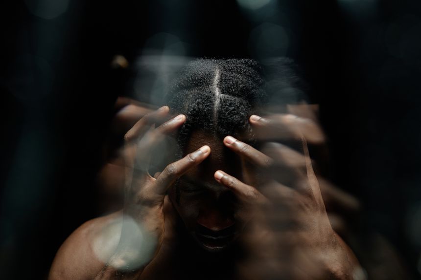 Abstract shot through glass with water running down of depressed Black man covering head with hands hiding face in deep shadow, casting from contrast top light in studio with black background