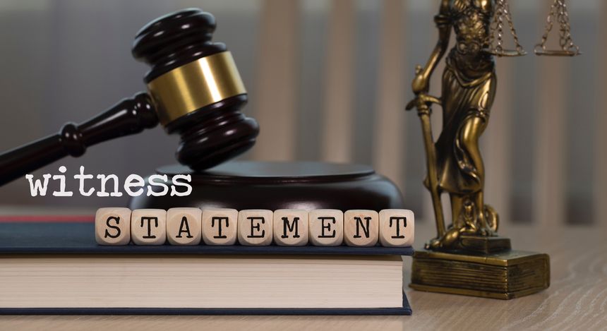 Word WITNESS STATEMENT composed of wooden dices. Wooden gavel and statue of Themis in the background. Closeup