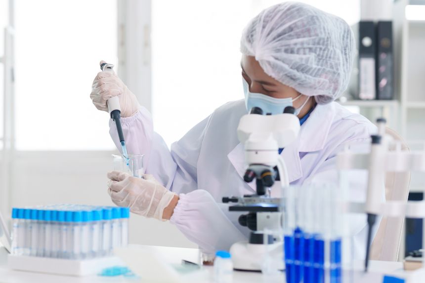 Dedicated Asian adult scientist performs laboratory experiment transferring liquid with pipette examining samples via microscope focused on scientific investigation.