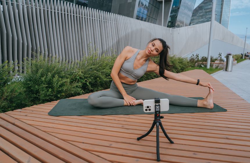 Woman stretching on a yoga mat outdoors with a camera on a tripod, modern architecture in the background