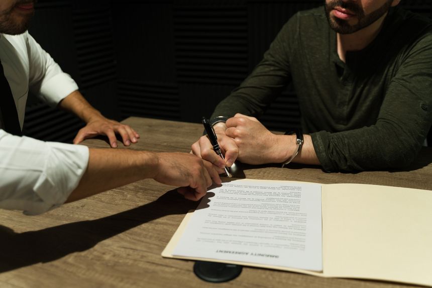 Cloaked in shadow, a prisoner signs an immunity deal during an interrogation with a detective