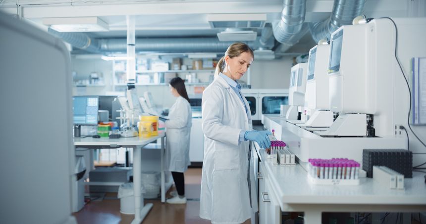 Biomedical Researchers Conduct Experiments in a Modern Laboratory. Female Scientist Preparing a Set of Samples in Test Tubes with Purple Caps. Colleague Analyzes Data on a Computer in the Background