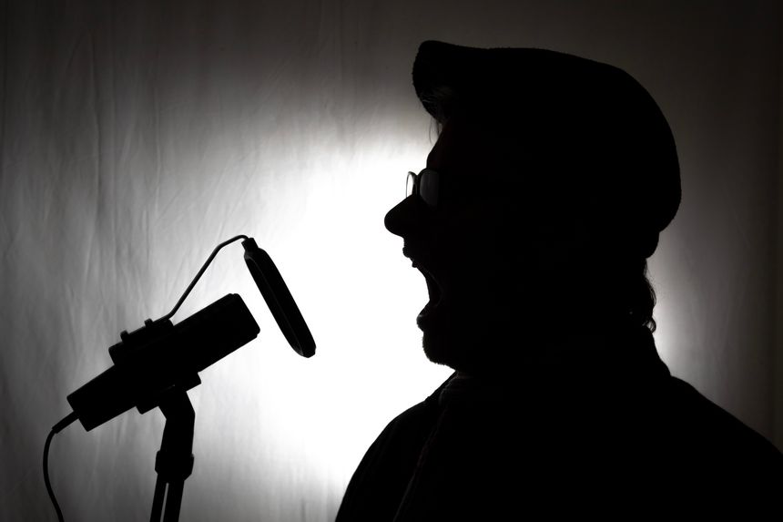 Voiceover artist silhouette shouting in a studio with microphone and pop filter wearing a hat against a black and white background