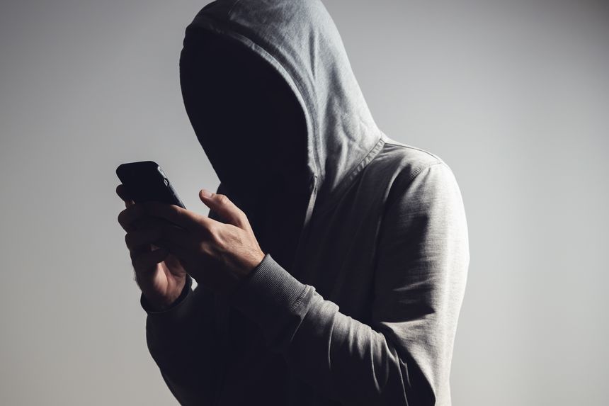 Man in hoodie using smartphone in dark shadow, face hidden in black, concept of anonymity, secrecy, cybercrime, or privacy. Selective focus.
