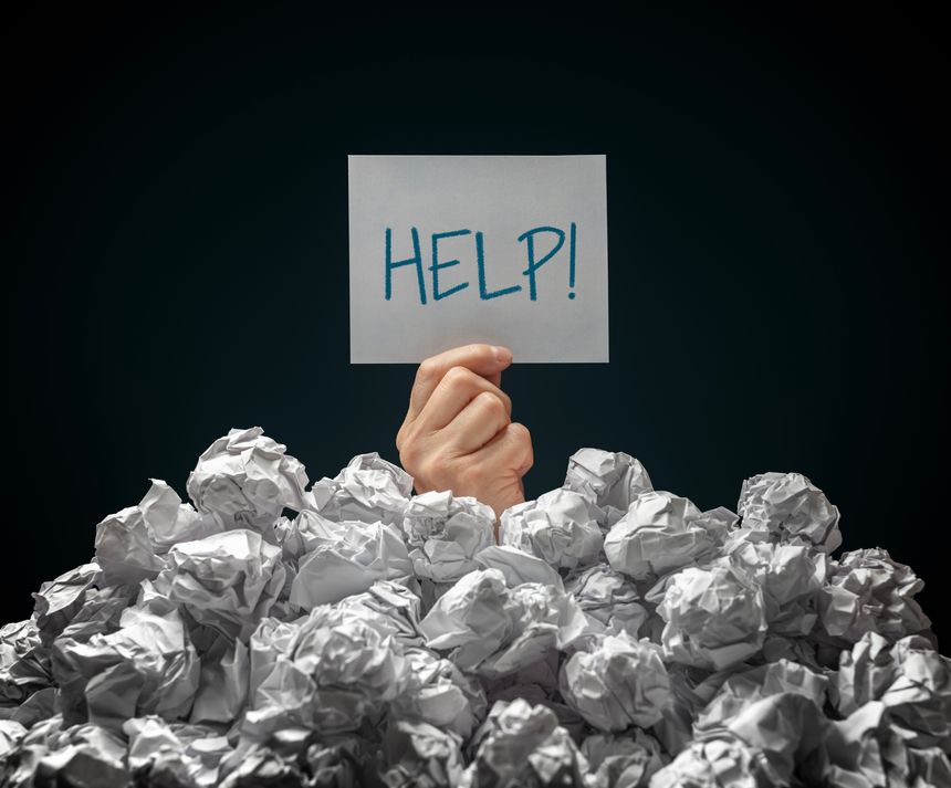 Businessman buried under crumpled pile of paperwork papers with a help sign with black background