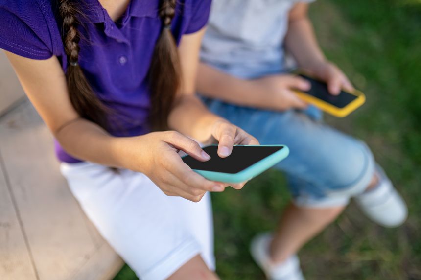 Concentration. Children in casual light clothes with smartphones sitting outdoors on summer day, no faces