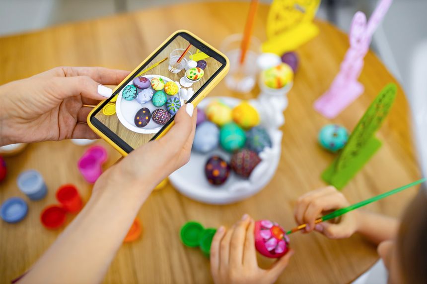 A mother and child are painting and decorating Easter eggs together on a wooden table, capturing the moment with a smartphone