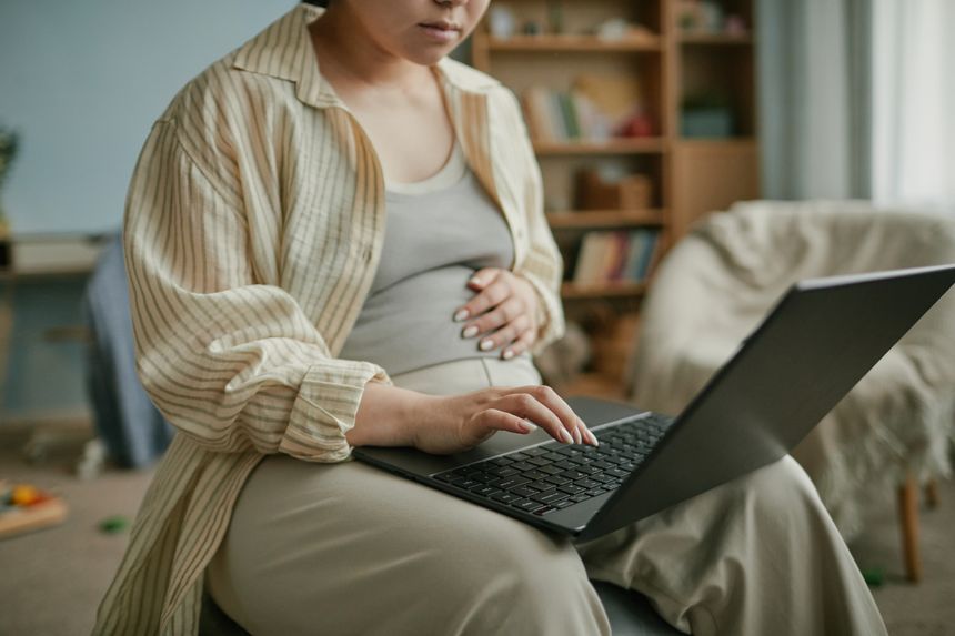 Pregnant Asian woman sitting indoors using laptop with one hand while gently touching belly, focusing on screen, casual home environment, preparing for motherhood, young adult