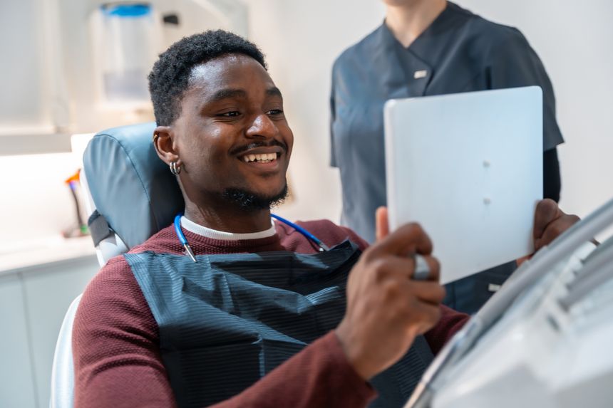 Cheerful young man is sitting in a dentist's chair, looking at a tablet held by a dental assistant, likely reviewing dental scan results