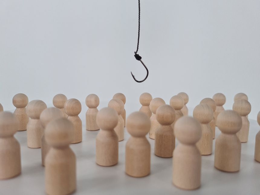 A single black fishing hook hangs from a thin string, suspended above a large group of simple wooden figures