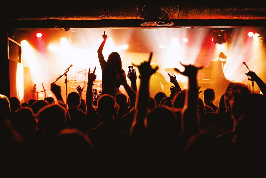 A girl sits on someoneâs shoulders in a concert crowd with her arm raised toward the bright stage. Around her, fans gesture and cheer during an intense live rock show.
