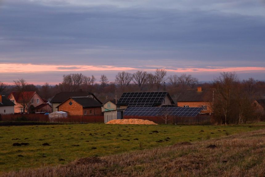 A panoramic view of a rural area at evening twilight under a pink and blue sky. Modern solar panels installed on rooftops and in yards stand out against the background of cozy private houses,