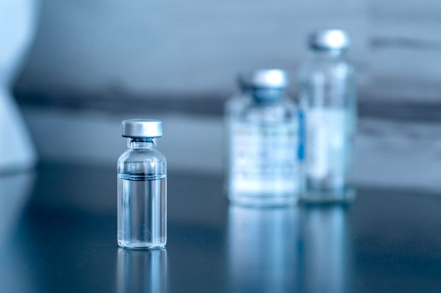 Transparent glass vials with metal caps. A close-up of several vials arranged on a reflective surface. The liquid contained in the vials is colorless and transparent. The lighting is soft and creates a blurred background effect, highlighting the vials in the foreground.