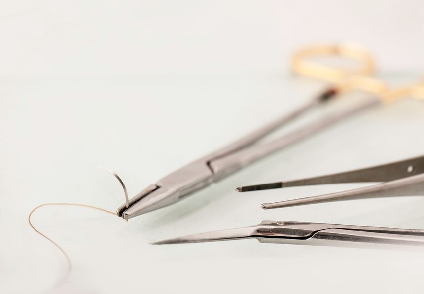 Color image of a set of surgical suturing instruments consisting of steel needle holder with suture and needle, forceps and scissor, resting on white surface