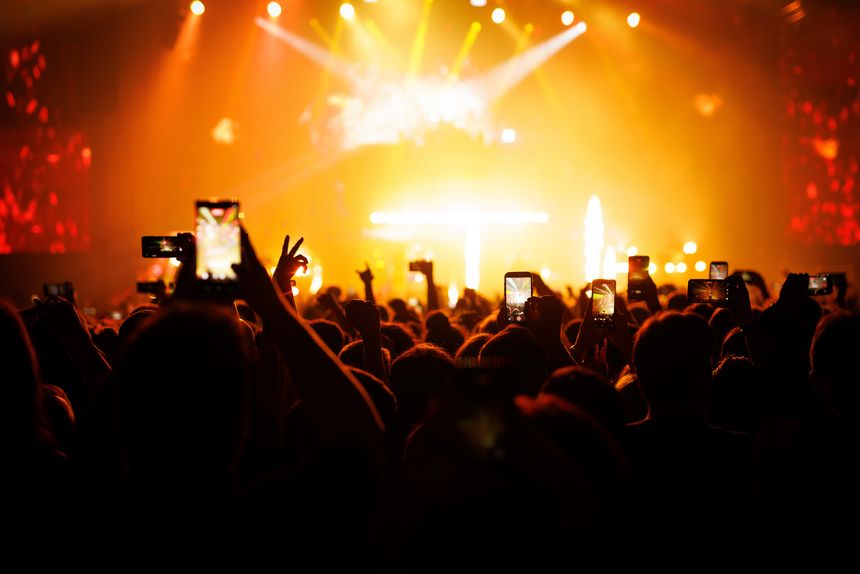 A massive audience captures a live music performance using multiple elevated smartphones. The entire scene is bathed in an intense, fiery orange glow from the overhead stage spotlights.