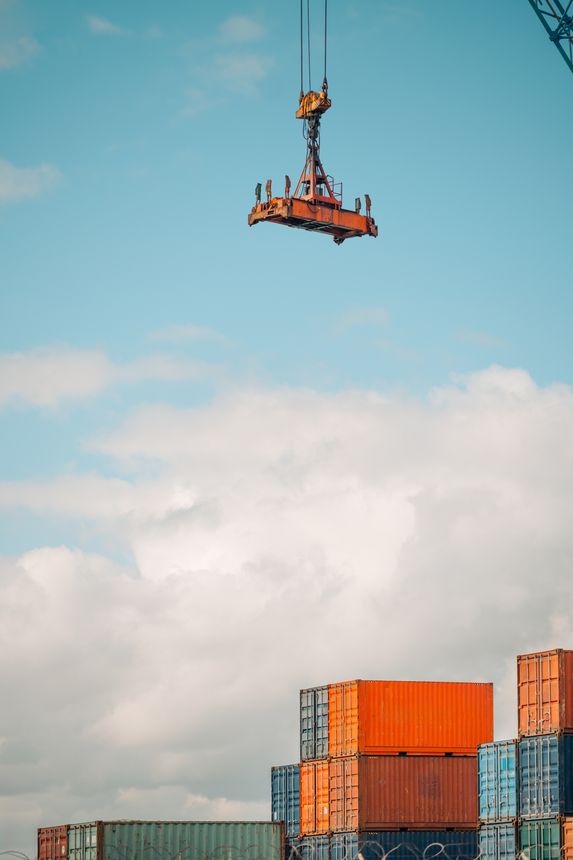 A large industrial crane hook and spreader lifting mechanism is suspended in the air against a bright blue sky with scattered clouds at a busy shipping port, above stacked cargo containers