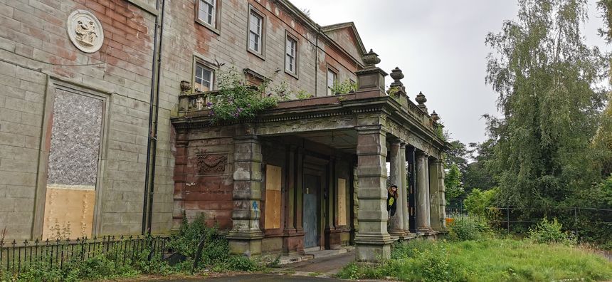 Derelict Woolton Hall building in Liverpool, Merseyside on 14th Aug 2021. Abandoned Stately Home Mansion in Merseyside, UK is boarded up in disrepair with broken windows and collapsing structure. Pillared porte-cochère car port area in front grounds of the estate showing porch doorway entrance to dilapidated home and overgrown gardens. Former house falling apart after years of neglect, arson and vandalism.