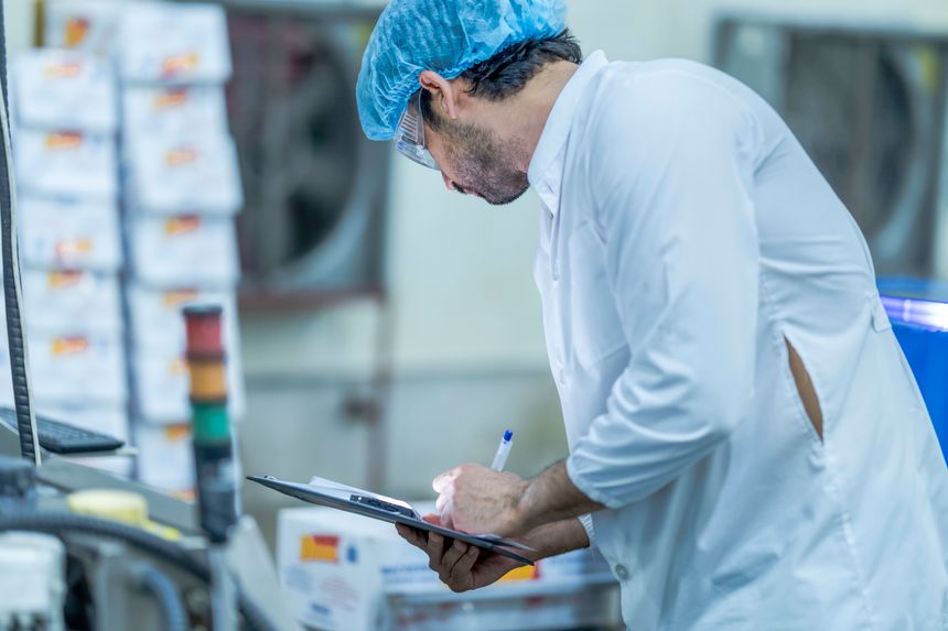 A food processing supervisor wearing protective gear and holding a clipboard, inspecting machinery on a production line, emphasizing quality control, safety, and efficiency in industrial operations.