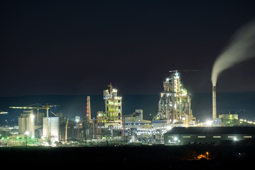High concrete structure of cement factory with tower crane and smoking chimney at night. Industrial production and air pollution concept.