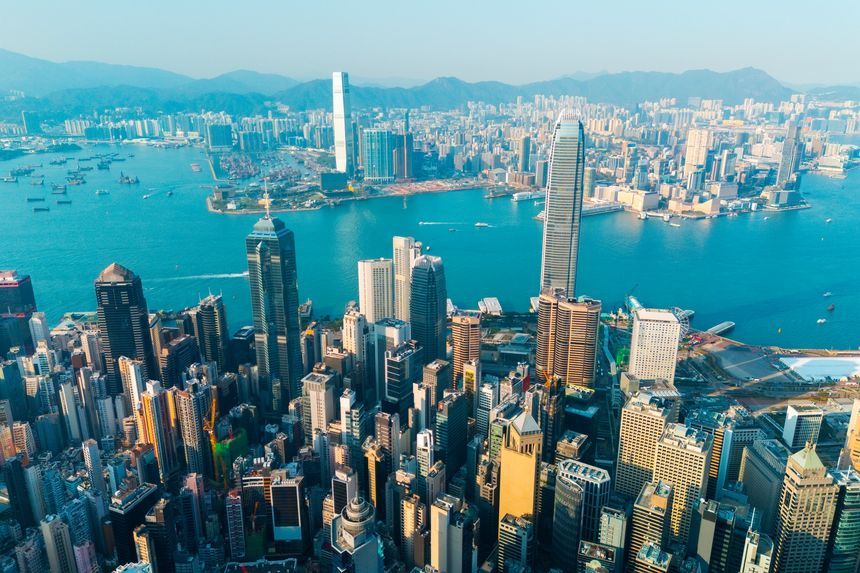 Numerous high-rise buildings dominate the urban landscape of Hong Kong. The blue waters of the harbor are dotted with boats and vessels, while the distant mountains provide a scenic backdrop.