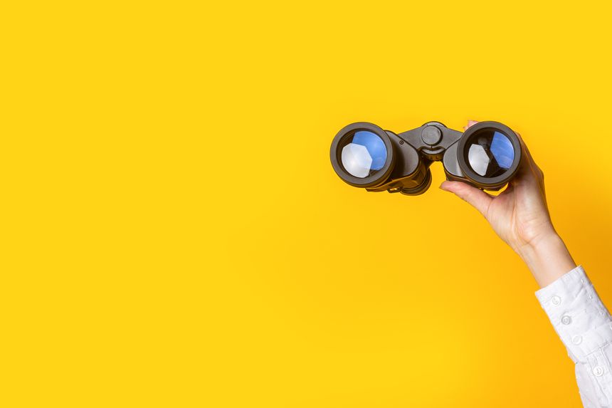 female hand holds black binoculars on a bright yellow background.