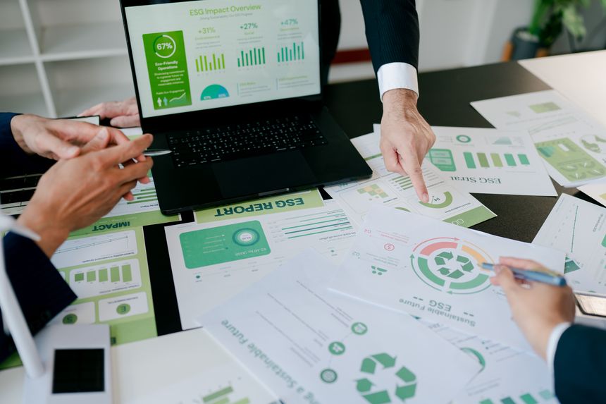 Business professionals' hands pointing at various esg reports and charts on a laptop and papers, discussing sustainable development goals and environmental friendly operations in a corporate setting