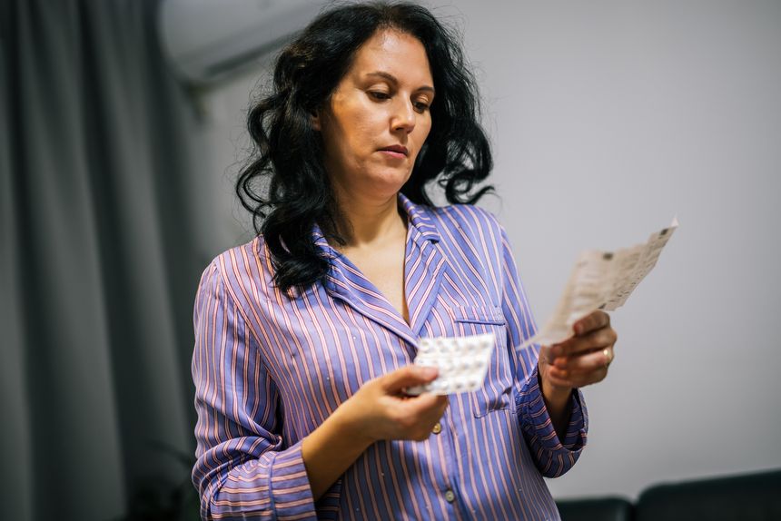 Middle-aged woman at home in pajamas carefully checking the instructions on a medicine leaflet before taking her medication.