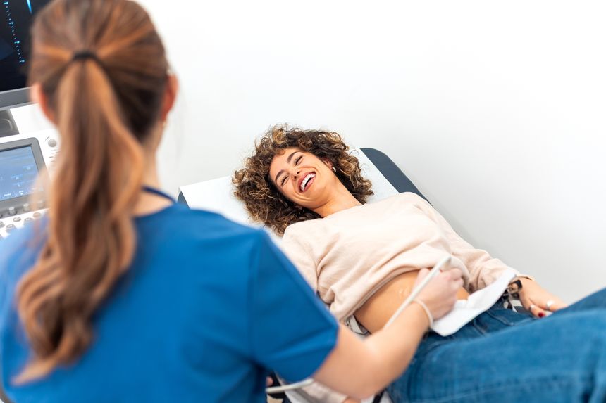 Happy pregnant woman laughing during ultrasound check in modern clinic. Healthcare and medicine concept. Female doctor screening patient belly with scanner for baby development or pregnancy test.